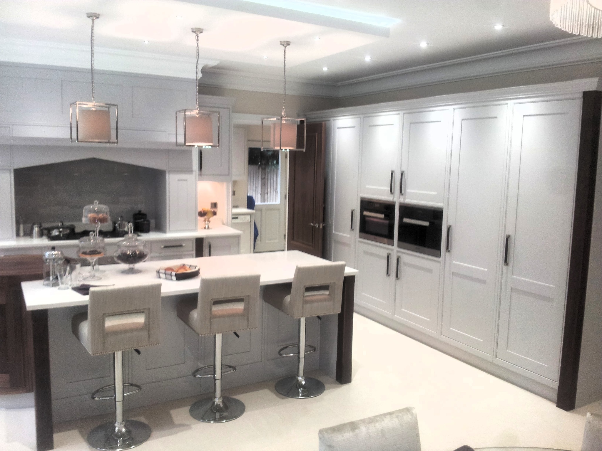 Elegancka, jasna kuchnia z wyspą i trzema hokerami. Zabudowa szafek pod sufit z wbudowanymi piekarnikami. Nad wyspą wiszą trzy lampy z abażurami.