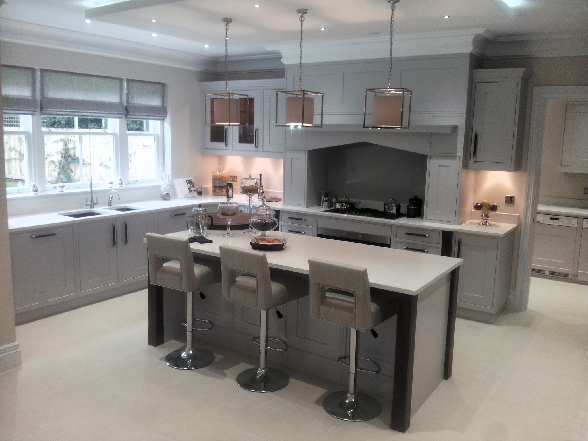 Jasna kuchnia na wymiar w stylu klasycznym: szare szafki z ciemnymi uchwytami, wyspa z białym blatem i hokerami, wiszące lampy w metalowych ramach. Okno z roletami rzymskimi.