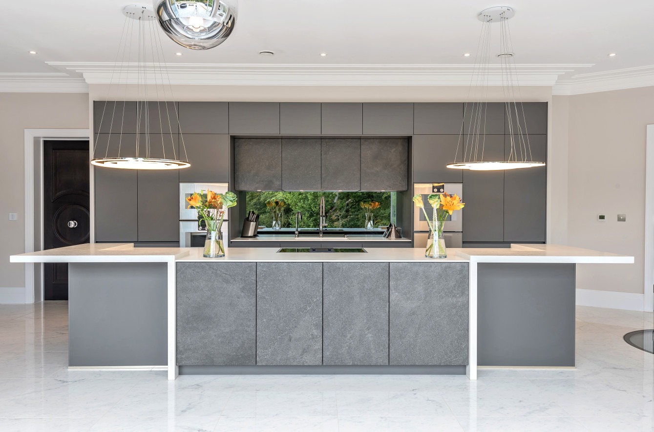 Nowoczesna, szara kuchnia z wyspą i blatem z białego konglomeratu. Nad wyspą wiszą okrągłe lampy. W tle widać zabudowę kuchenną z oknem i kwiatami.