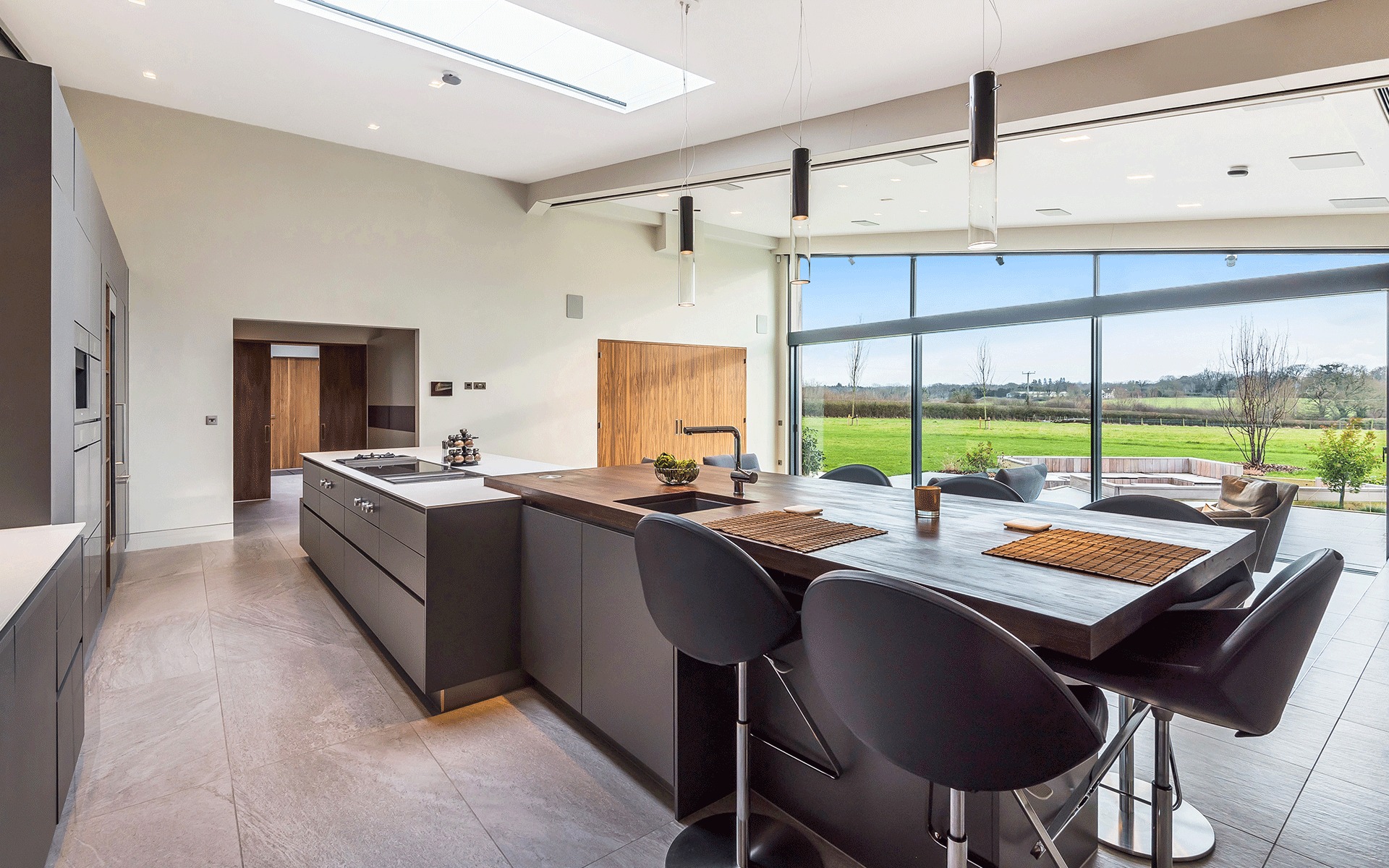 Nowoczesna kuchnia z wyspą i dużym oknem z widokiem na zieleń. Ciemne, matowe fronty szafek kontrastują z drewnianym blatem. Minimalistyczne oświetlenie.