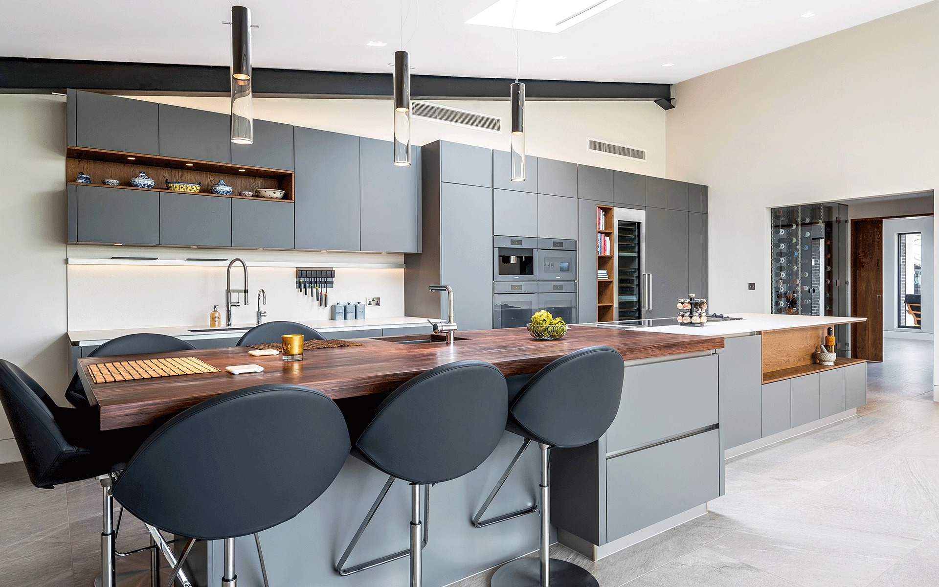 Nowoczesna kuchnia z wyspą i drewnianym blatem, szare szafki z oświetleniem LED, wiszące lampy. Nietypowy układ szafek górnych z otwartą półką z dekoracjami.