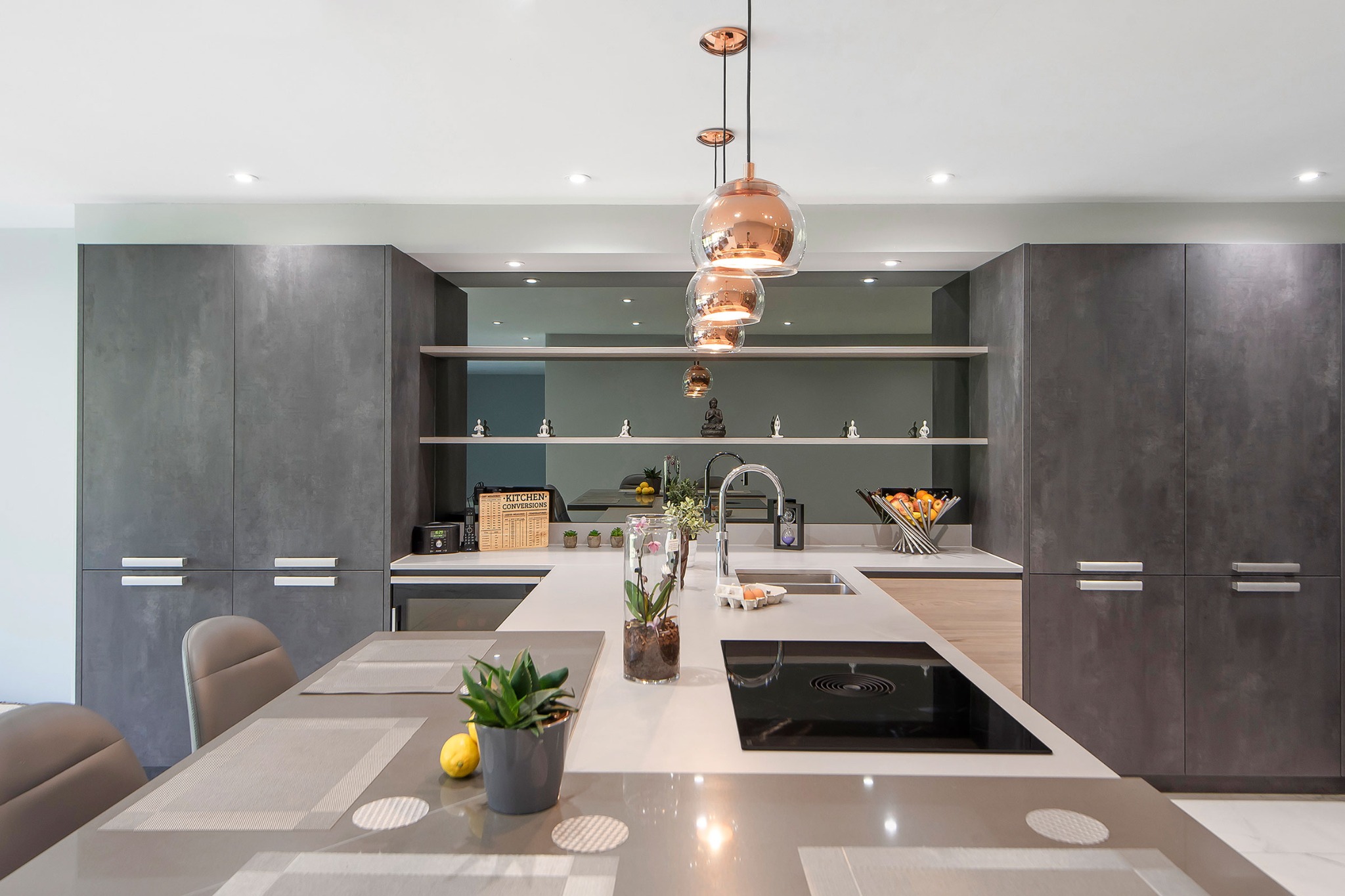 Nowoczesna kuchnia z szarymi szafkami, wyspą z płytą indukcyjną i złotymi lampami wiszącymi. Na półkach dekoracje.