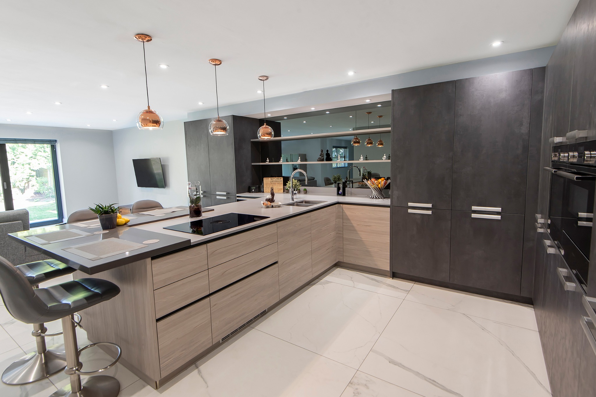 Nowoczesna kuchnia z wyspą, fronty w odcieniach szarości i beżu, biała marmurowa podłoga. Nad wyspą wiszą miedziane lampy. W tle widoczne okno i salon.