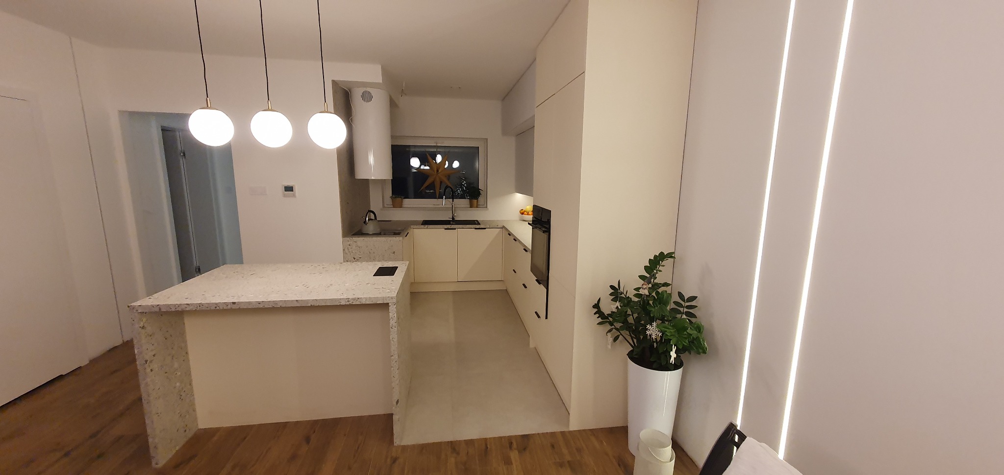 Nowoczesna, jasna kuchnia z wyspą wykończoną blatem z lastryko. Minimalistyczne oświetlenie, gładkie fronty szafek i dekoracje świąteczne w oknie nadają wnętrzu przytulny charakter.