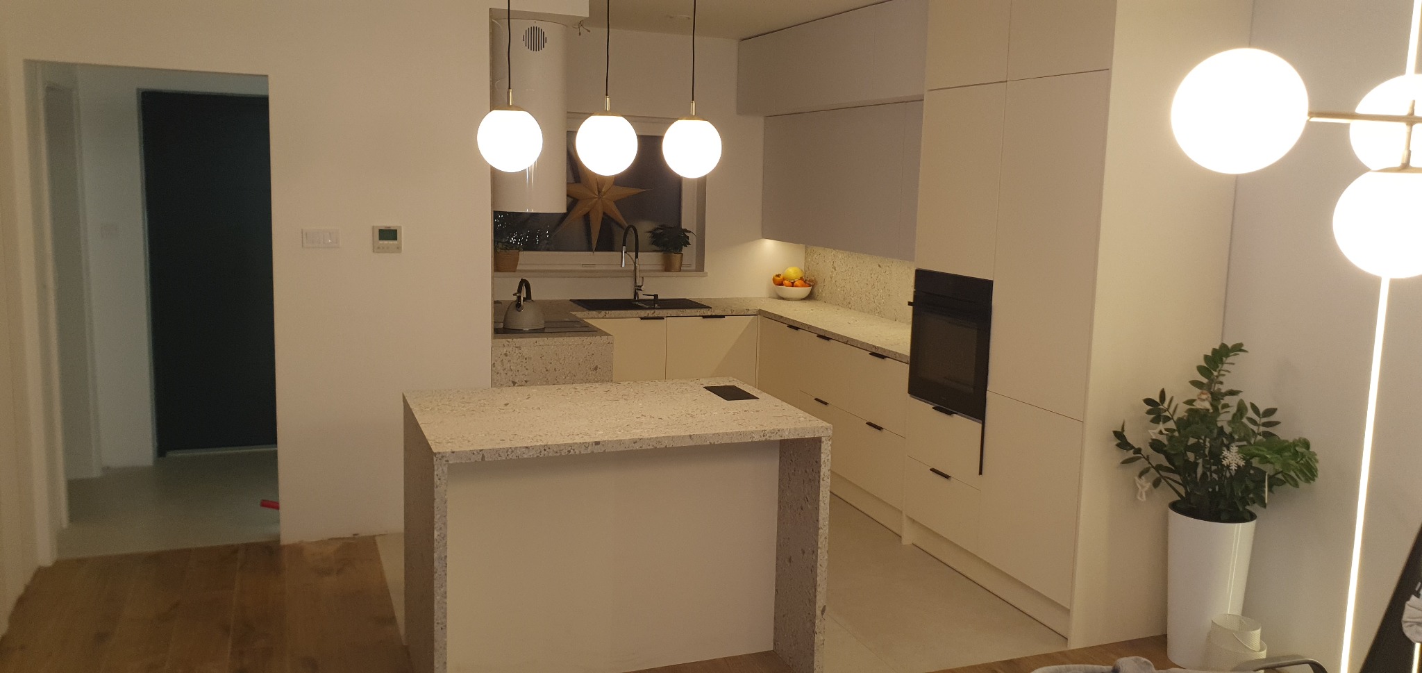 Nowoczesna kuchnia z wyspą, jasne fronty szafek, blat z kamienia kompozytowego, oświetlenie punktowe i wiszące lampy kuliste. Minimalistyczny design.