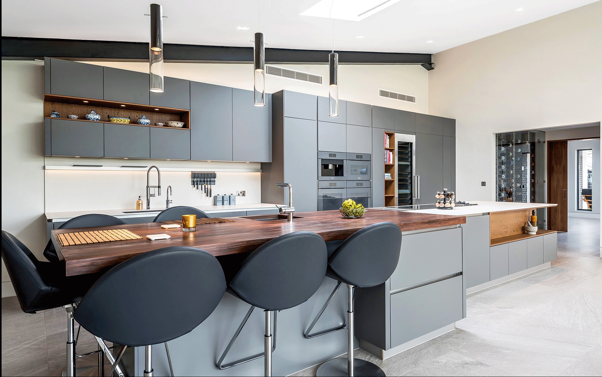 Nowoczesna kuchnia z szarymi szafkami, drewnianym blatem wyspy i designerskimi lampami. Otwarta przestrzeń, minimalistyczny styl. Widoczne sprzęty AGD i dekoracje.