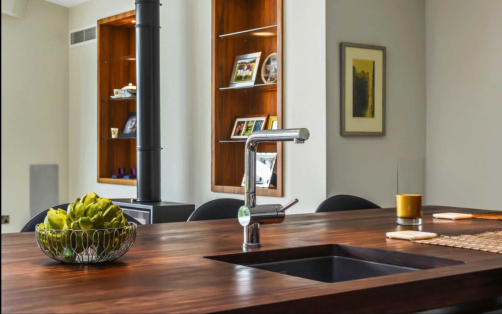 Nowoczesna kuchnia z blatem z ciemnego drewna i chromowaną baterią. W tle półki z dekoracjami i piec wolnostojący. Minimalistyczny design.