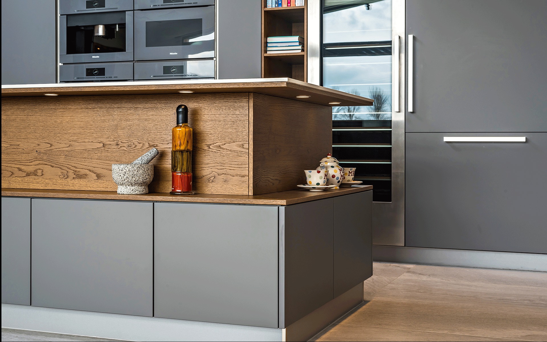 Nowoczesna kuchnia z szarymi szafkami i drewnianym blatem, oświetleniem LED. Na blacie moździerz, butelka z marynowanymi warzywami i zestaw do herbaty.