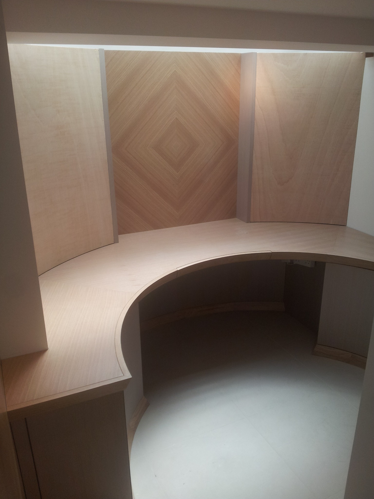 Biało-drewniana, zakrzywiona zabudowa biurowa z biurkiem i szafkami, z dekoracyjnym panelem ściennym w romby, podświetlona od góry.