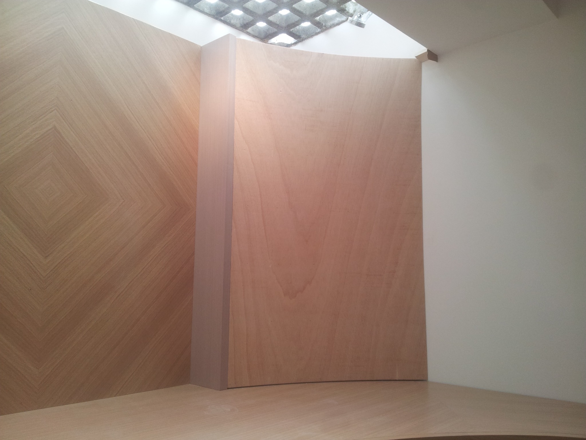 Narożnik z jasnego drewna z dekoracyjnym wzorem na ścianie i zakrzywionym elementem, oświetlony przez kratownicowy sufit.