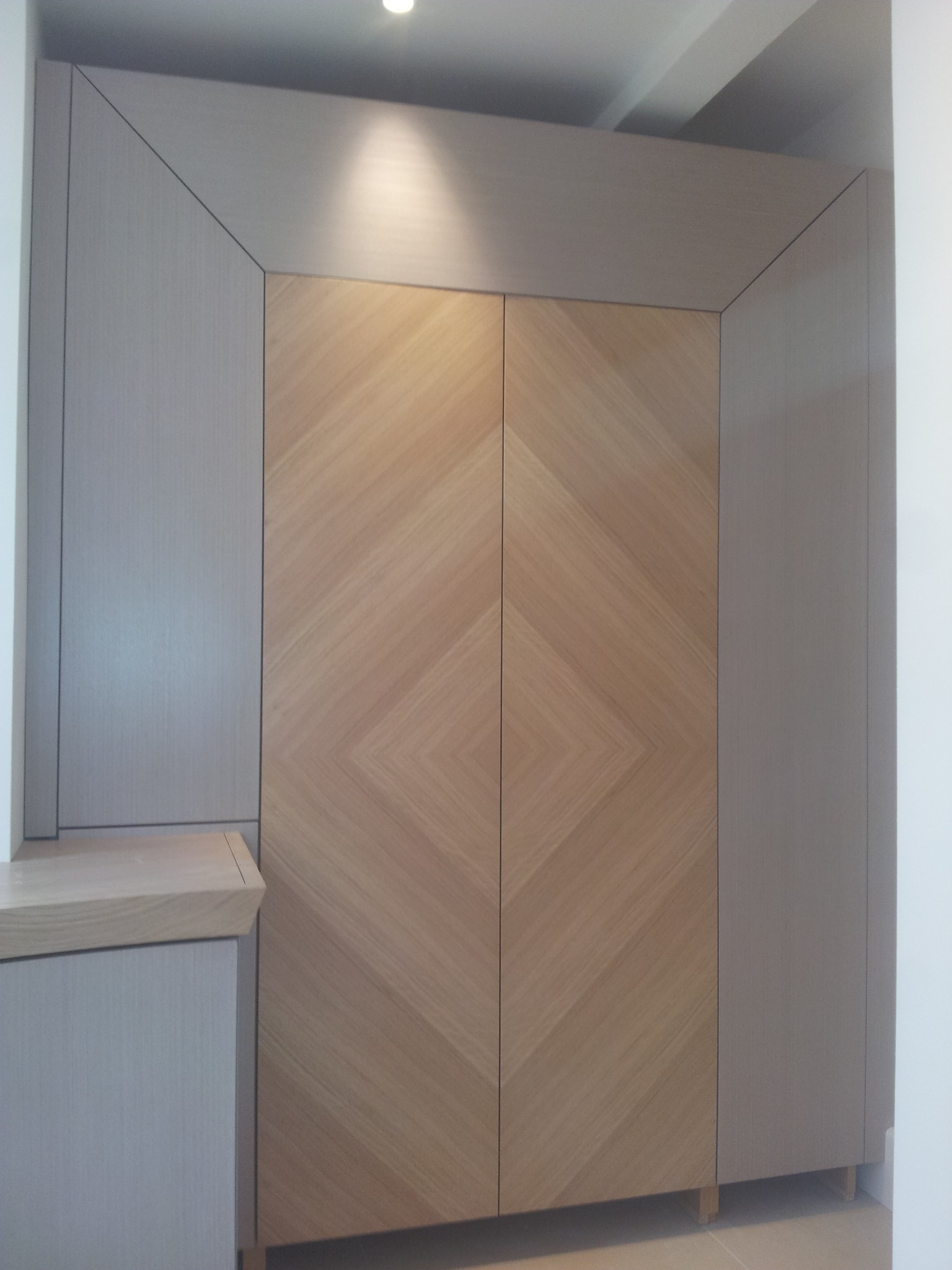 Dwudrzwiowa szafa wnękowa z drewnianymi frontami ułożonymi w jodełkę, otoczona szarymi panelami. Górne podświetlenie podkreśla teksturę drewna i geometryczny wzór.