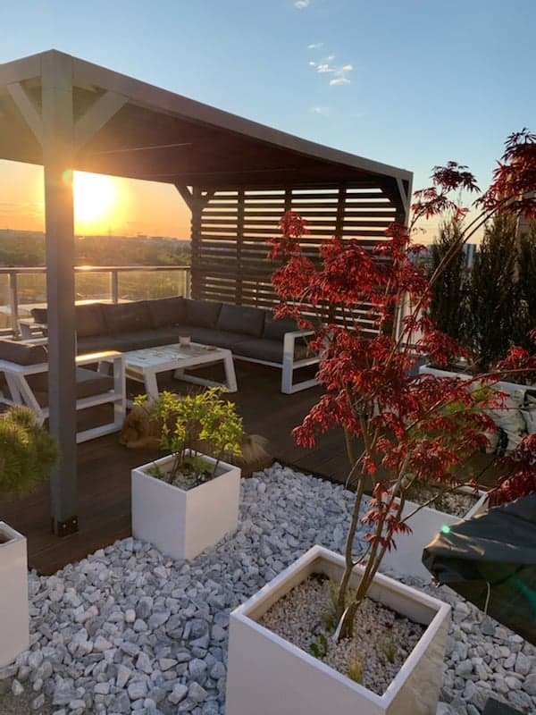 Nowoczesny taras na dachu z drewnianą podłogą, pergolą z poziomymi listwami i meblami wypoczynkowymi; widok na zachód słońca.