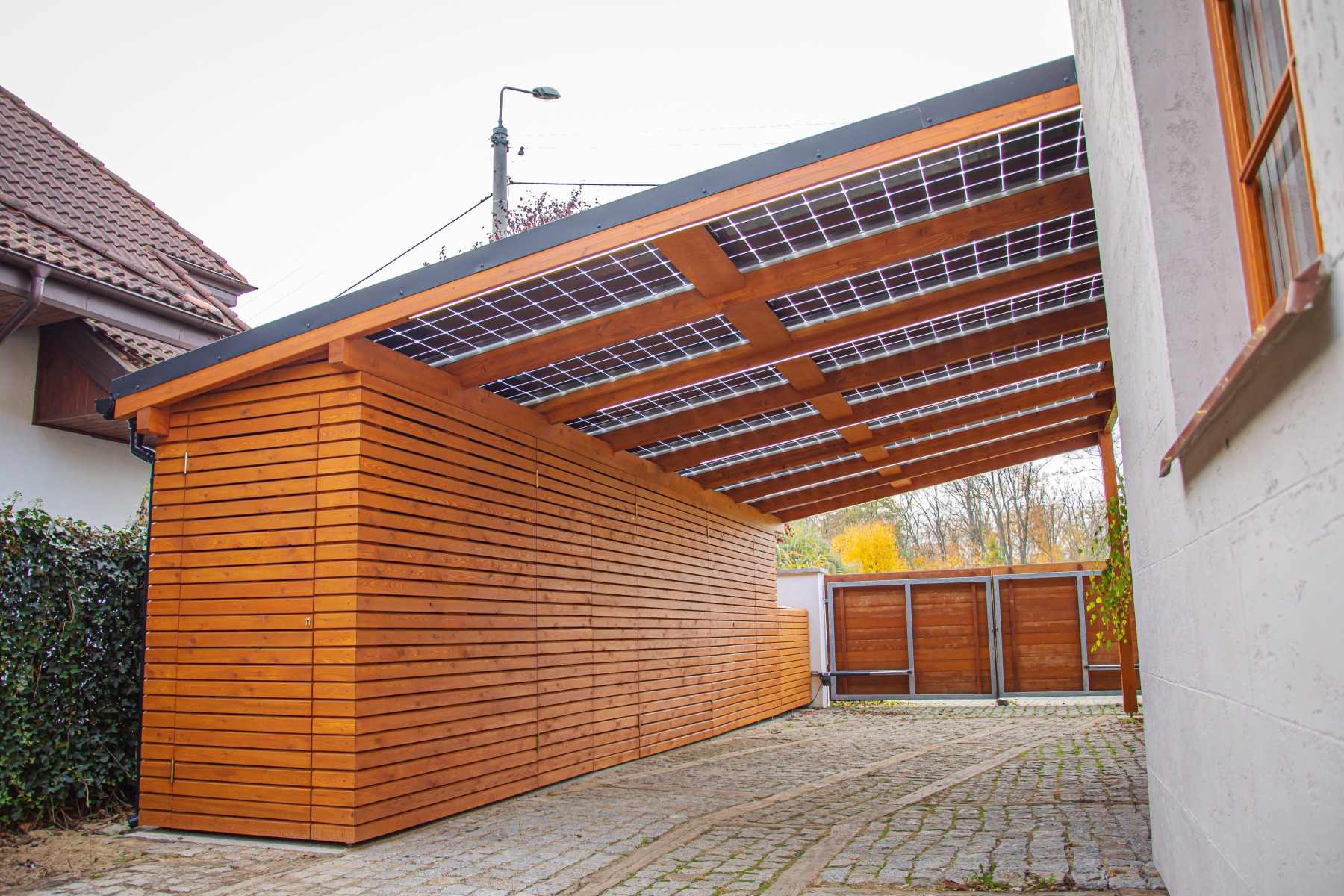 Nowoczesna wiata garażowa z drewna z panelami słonecznymi na dachu, widok na brukowany podjazd i bramę w tle.
