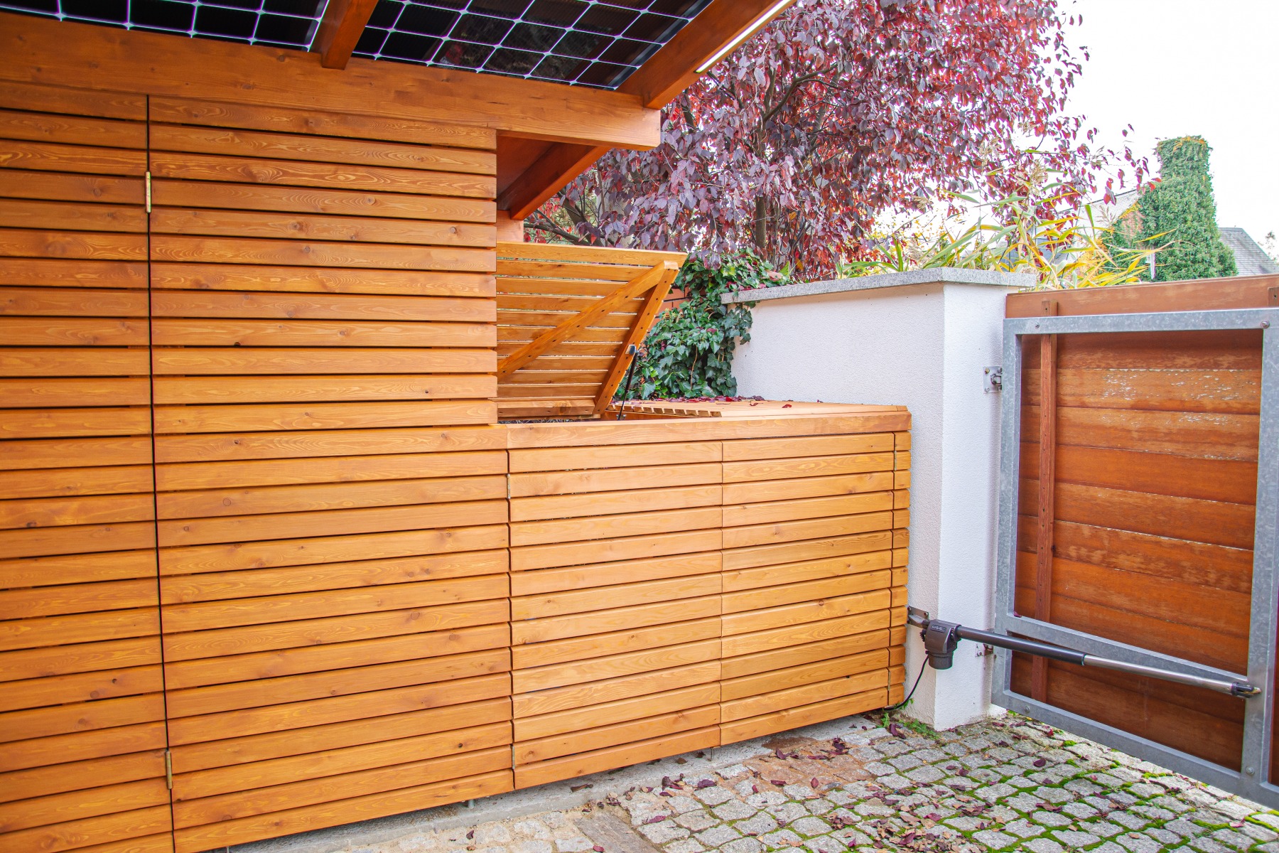 Drewniana zabudowa tarasu z otwieranym schowkiem, widoczna brama z automatycznym napędem i kostka brukowa na podłożu.
