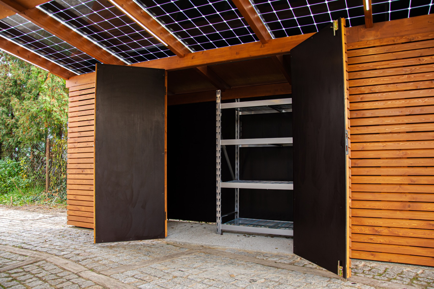 Drewniana konstrukcja z panelami słonecznymi na dachu i otwartymi, ciemnymi drzwiami, ukazującymi metalowy regał w środku.