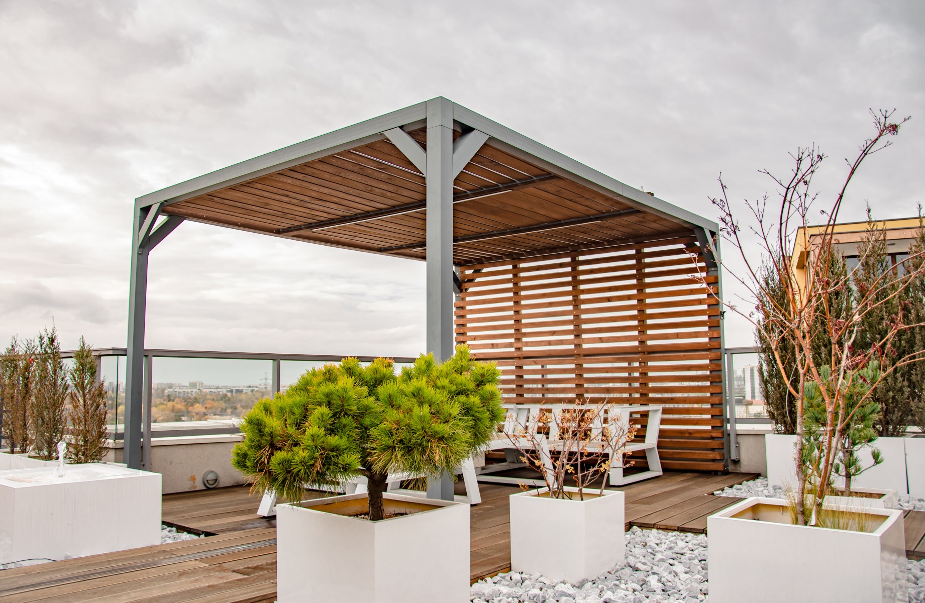Metalowa pergola z drewnianym zadaszeniem i ażurową ścianą na tarasie z drewnianą podłogą, otoczona białymi donicami z roślinami i białym żwirem, widok na miasto w tle.