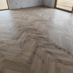 Andrzej Jezierski - Jasne panele ułożone w jodełkę na podłodze w pokoju z dużymi oknami. Ściany w stanie surowym, przygotowane do dalszych prac wykończeniowych. Widoczne plamy na panelach.