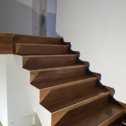 Wykończone drewniane schody z białymi podstopnicami, zabezpieczone niebieską taśmą malarską przy ścianie, widoczne antypoślizgowe paski.