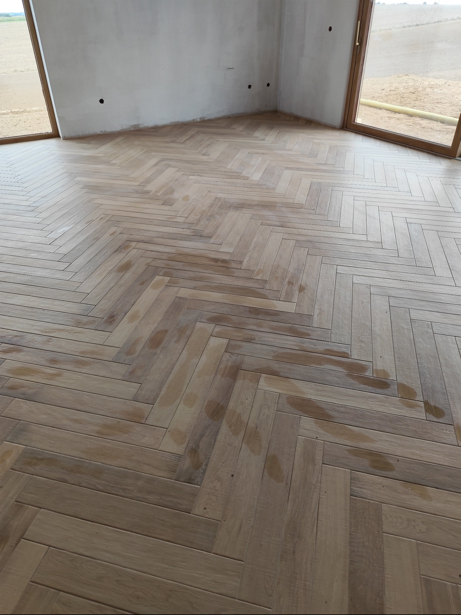 Jasne panele ułożone w jodełkę na podłodze w pokoju z dużymi oknami. Ściany w stanie surowym, przygotowane do dalszych prac wykończeniowych. Widoczne plamy na panelach.