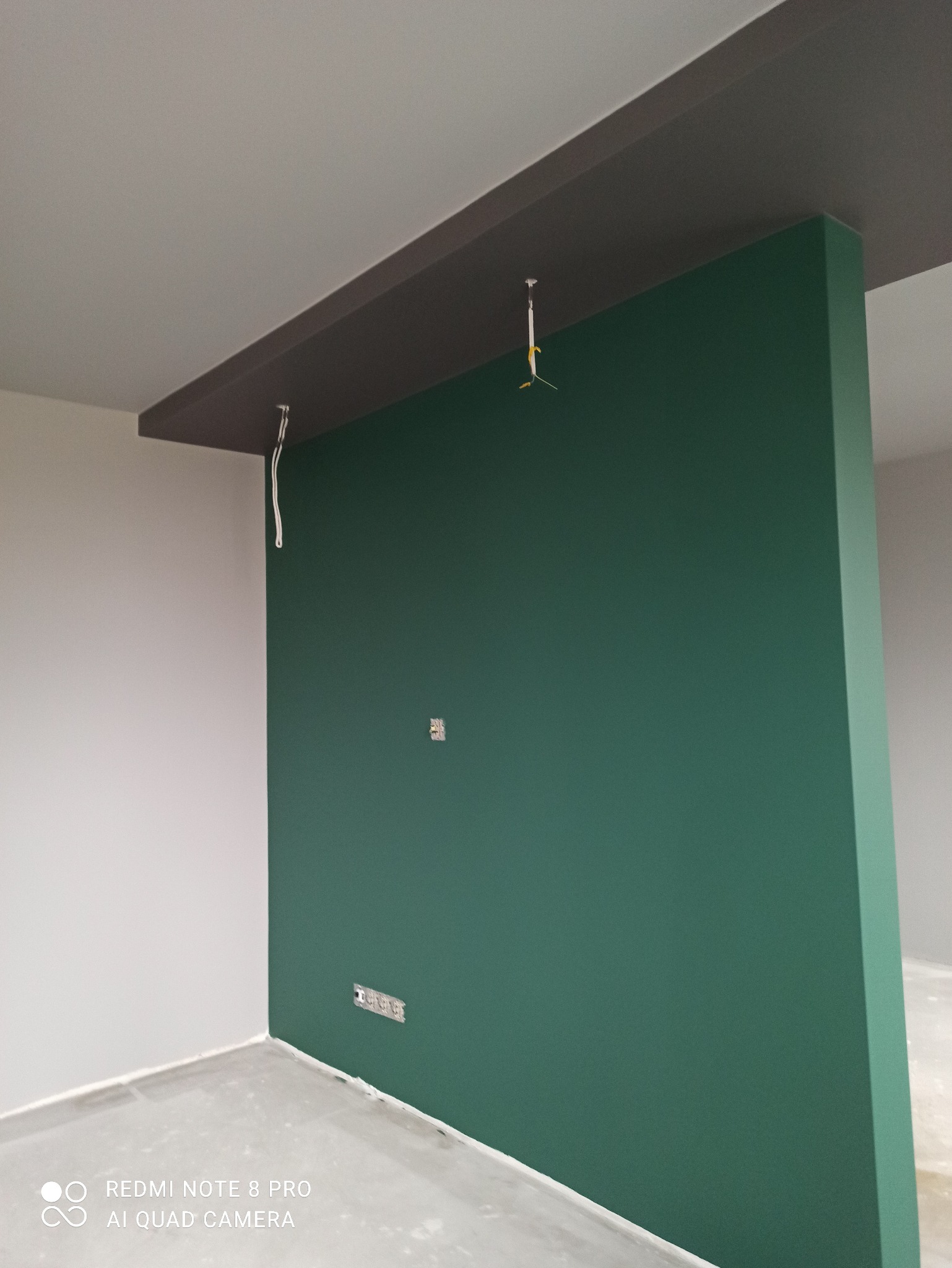 Ściana działowa pomalowana na zielono, z widocznymi gniazdkami elektrycznymi i wystającymi kablami oświetleniowymi. Sufit pomalowany na szaro. Świeżo wykończone wnętrze.