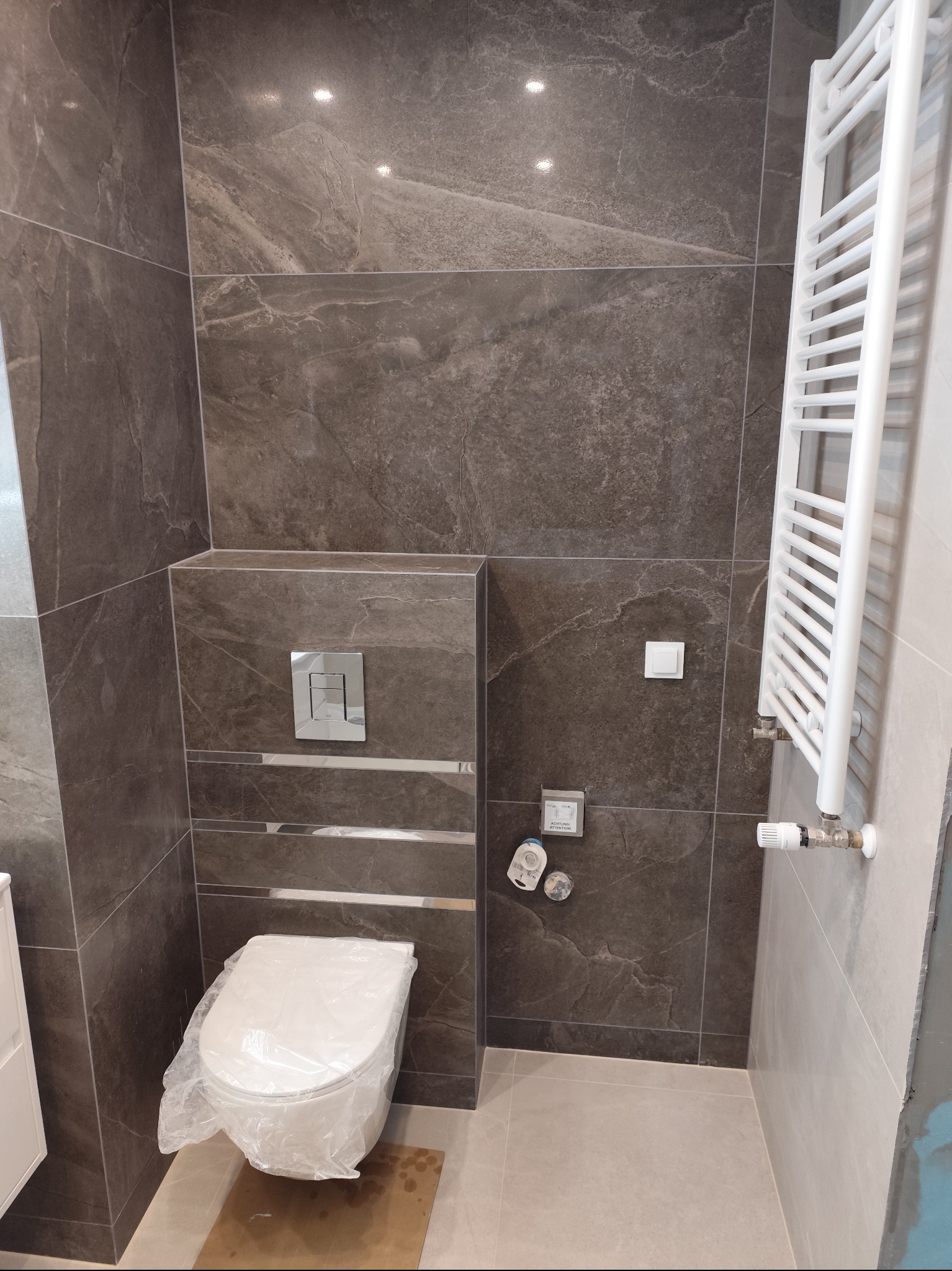 Wykończona łazienka z brązowymi płytkami imitującymi kamień, biały grzejnik drabinkowy i toaleta zabezpieczona folią. Nowoczesny design z chromowanymi akcentami.