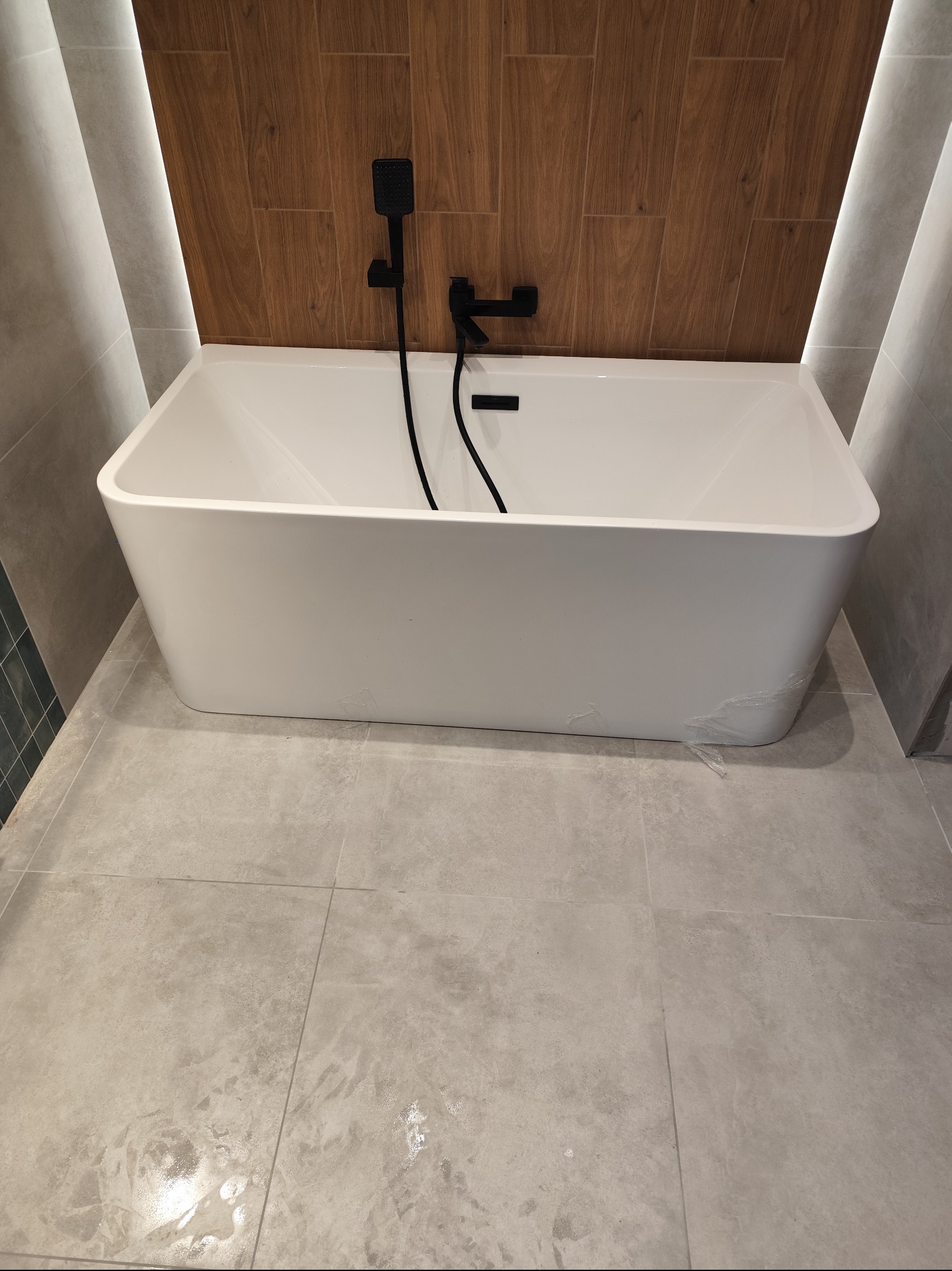 Nowoczesna, wolnostojąca wanna z czarną armaturą na tle drewnopodobnych płytek i szarych płytek podłogowych. Minimalistyczny design łazienki z subtelnym oświetleniem.