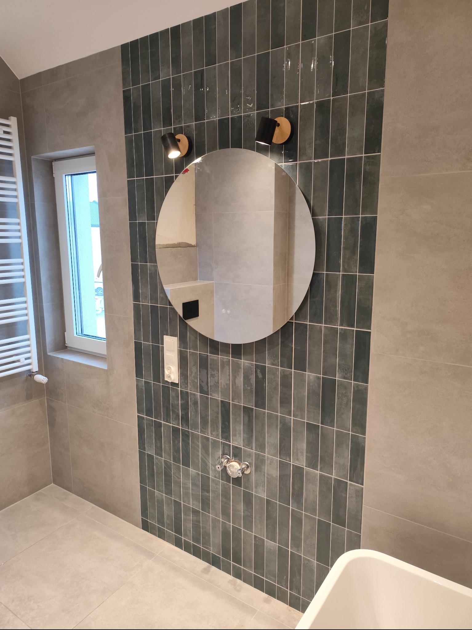 Wykończona łazienka w Iwaniskach z zielonymi, wąskimi płytkami na ścianie, okrągłym lustrem i nowoczesnym oświetleniem. Minimalistyczny design i jasne, stonowane kolory.