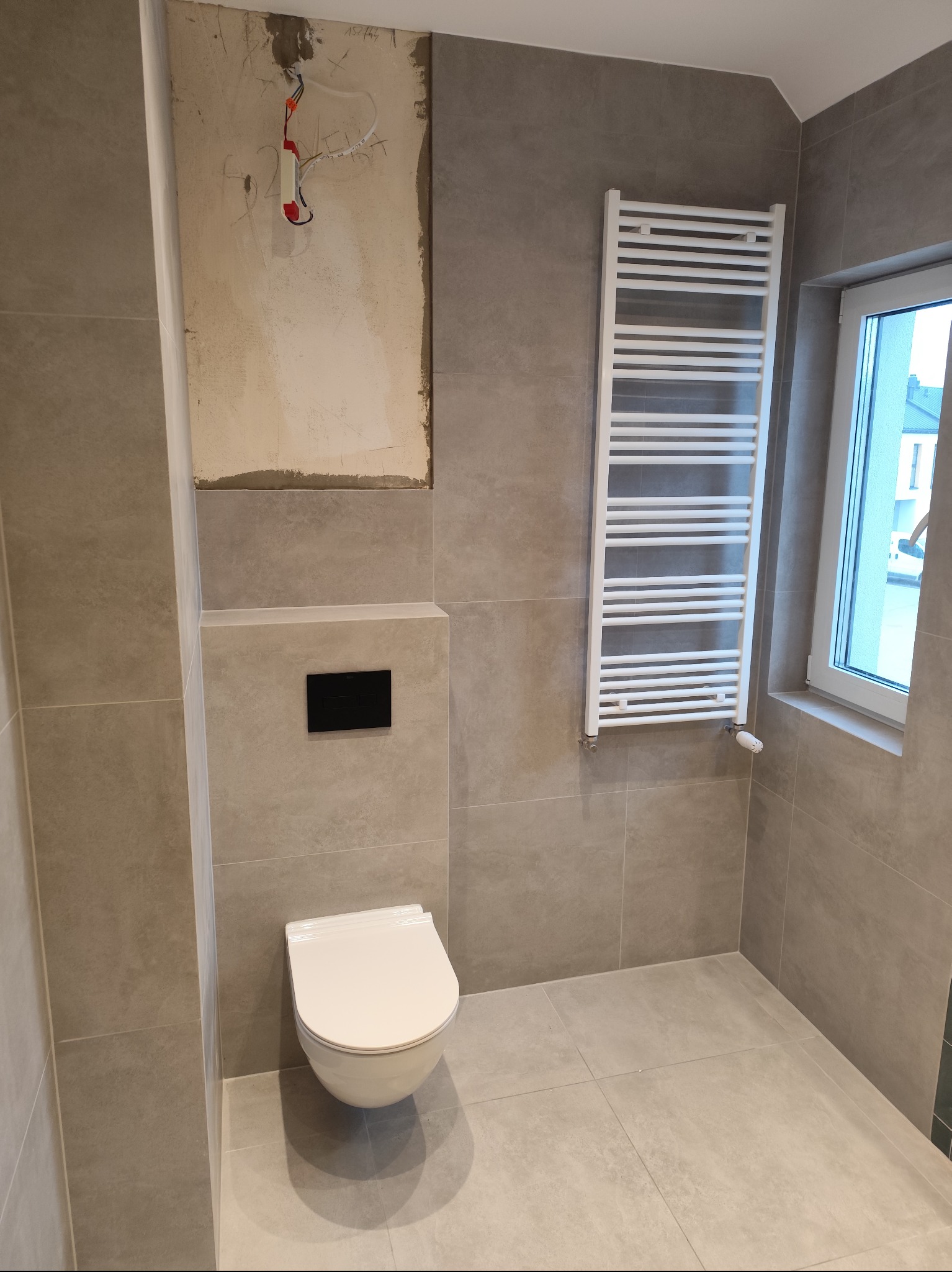 Wykończona łazienka w szarościach z podwieszaną toaletą, grzejnikiem drabinkowym i fragmentem ściany z widoczną instalacją elektryczną przed wykończeniem.