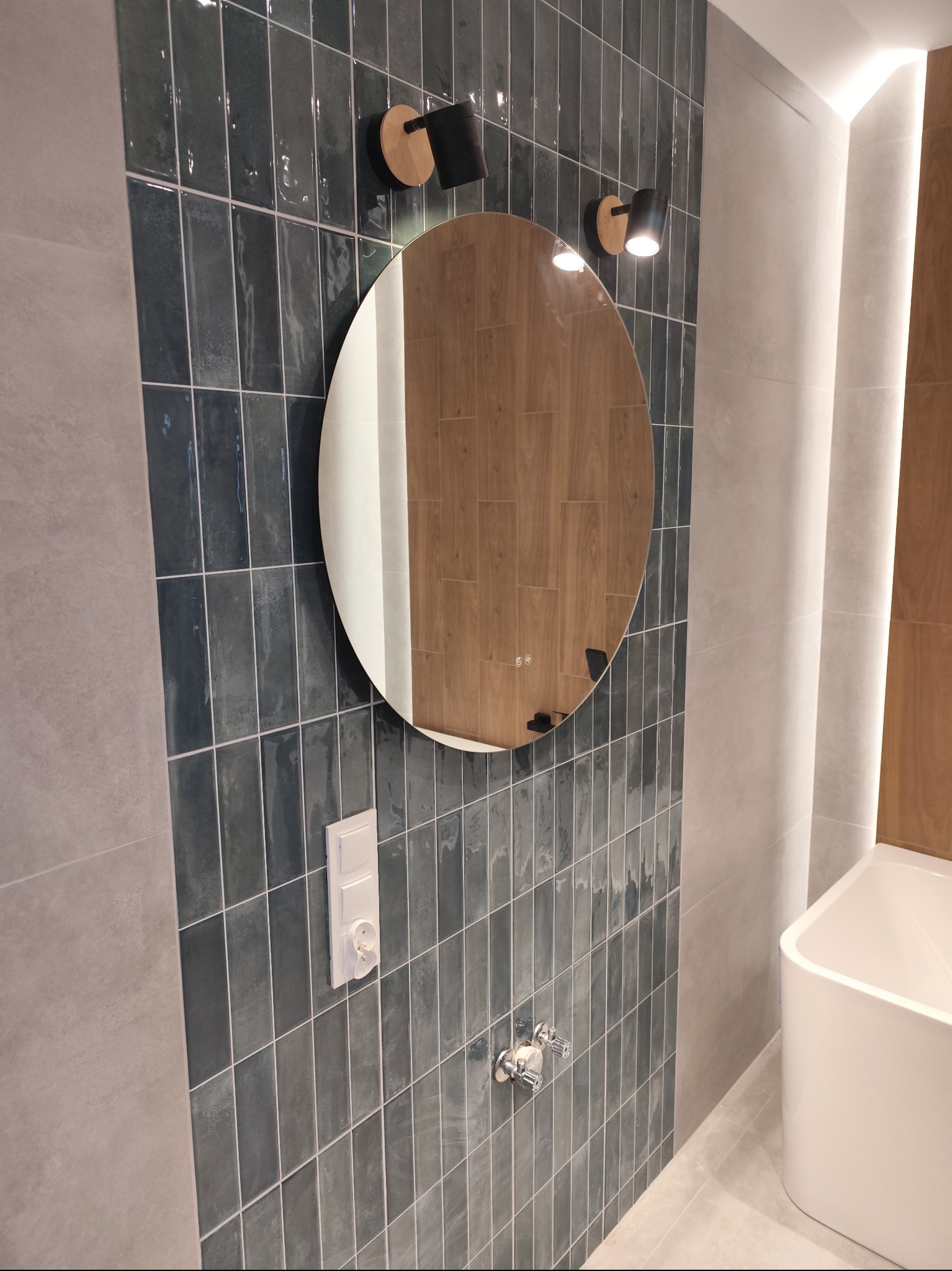 Błyszczące, pionowe, szmaragdowe płytki w łazience z okrągłym lustrem i armaturą. Widoczny fragment wanny i gniazdko elektryczne. Nowoczesny design.