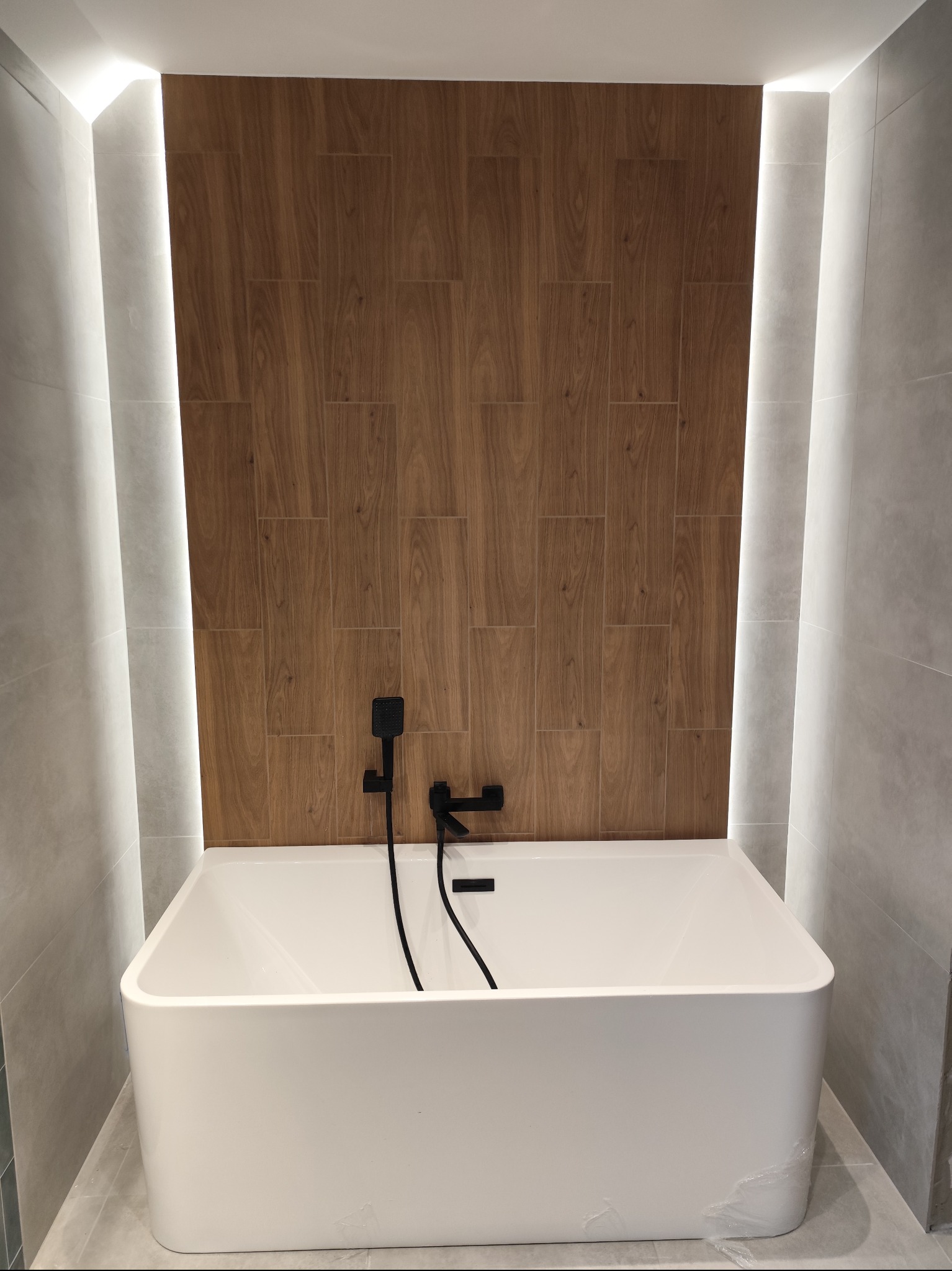 Nowoczesna łazienka z białą wanną, drewnopodobnymi płytkami na ścianie i subtelnym oświetleniem LED. Czarna armatura dodaje elegancji. Minimalistyczny design.