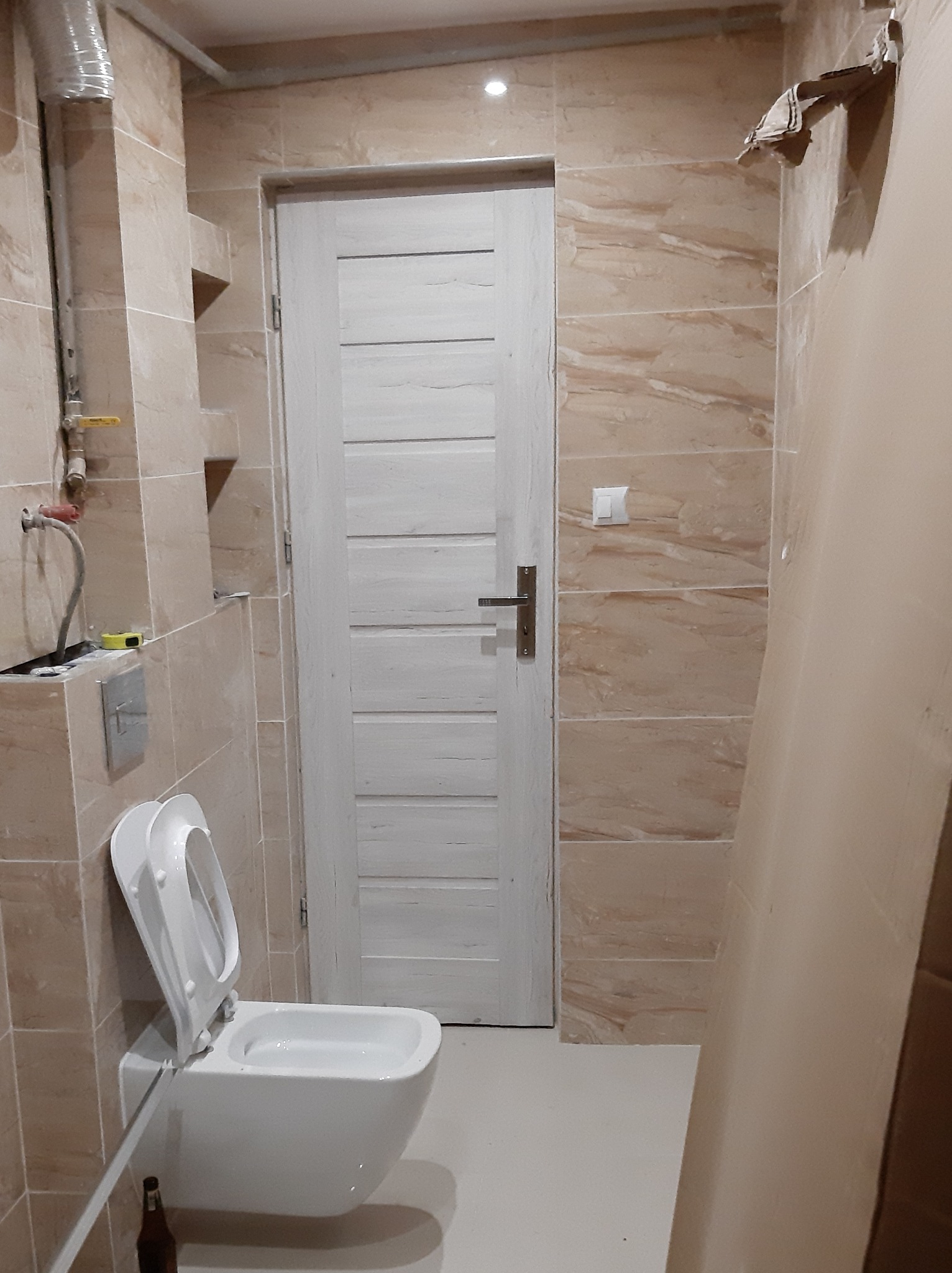 Wisząca toaleta w łazience z beżowymi płytkami imitującymi marmur, białe drzwi z poziomymi panelami i fragment instalacji wodno-kanalizacyjnej w rogu.