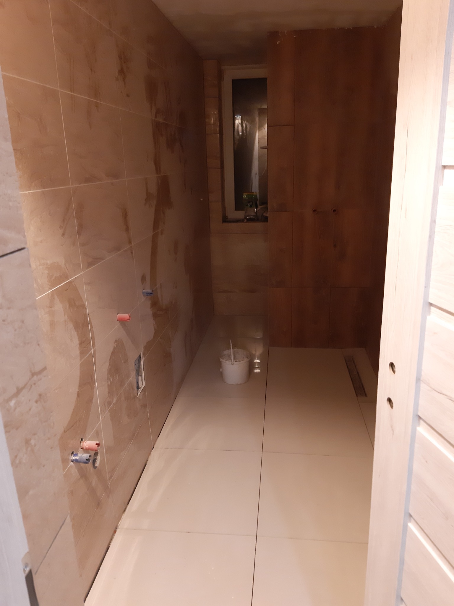 Łazienka w trakcie układania płytek, widoczne przyłącza wodne, wiadro i odpływ liniowy w podłodze.