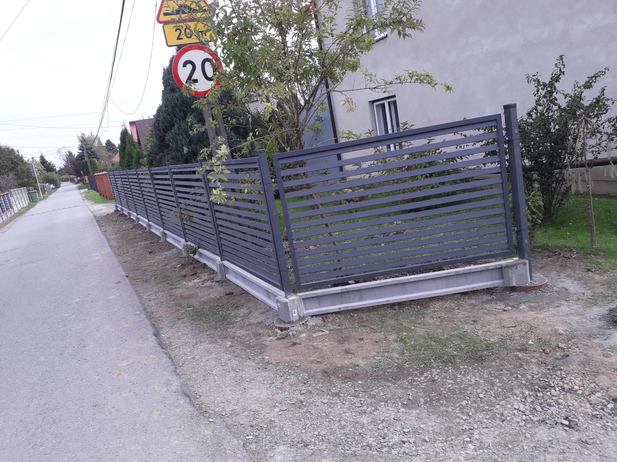 Nowoczesne, poziome, szare ogrodzenie panelowe z betonową podmurówką, widoczne od strony ulicy z znakiem ograniczenia prędkości do 20 km/h.