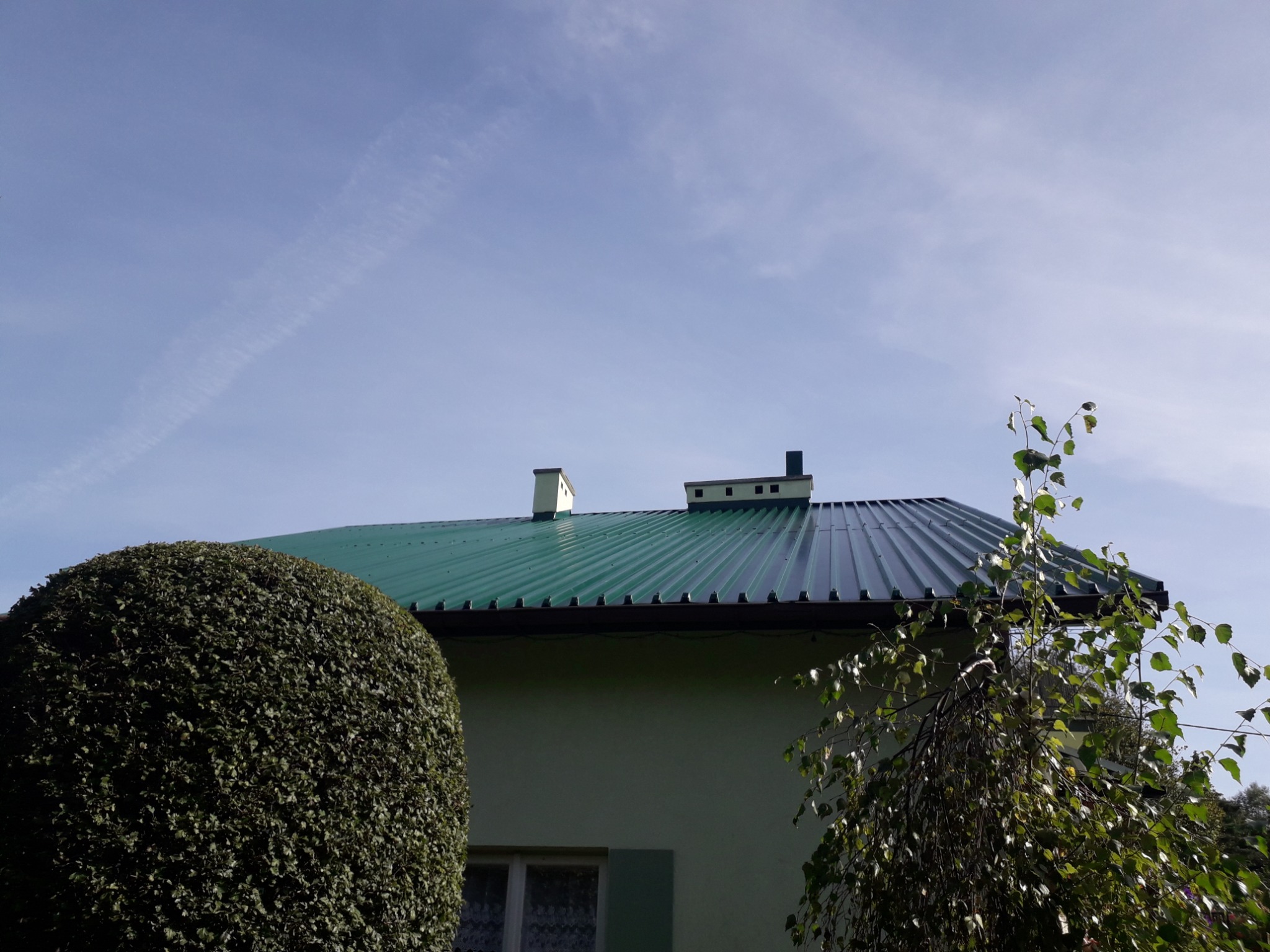 Zielony, blaszany dach domu jednorodzinnego z dwoma kominami, widoczny z poziomu ogrodu, na tle błękitnego nieba.
