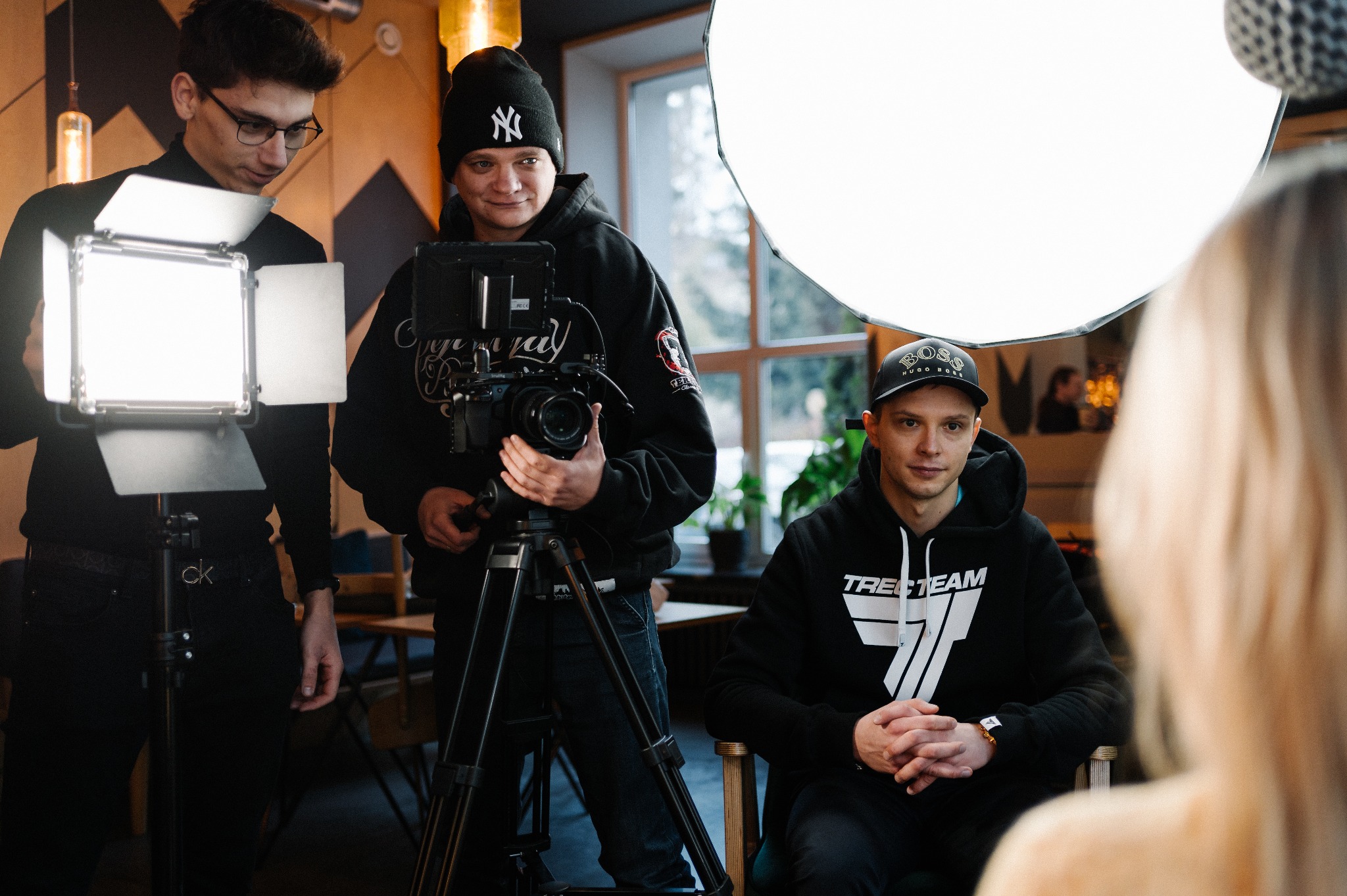 Ekipa filmowa podczas nagrywania ujęcia z oświetleniem LED i softboxem, operator kamery z czapką New York Yankees filmuje osobę siedzącą na krześle.