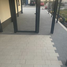 Firma Handlowo Usługowa Paweł Kulesza - Ułożona kostka brukowa w dwóch odcieniach szarości pod zadaszeniem, z widocznym fragmentem układania i pracującą osobą w tle, w otoczeniu domów.