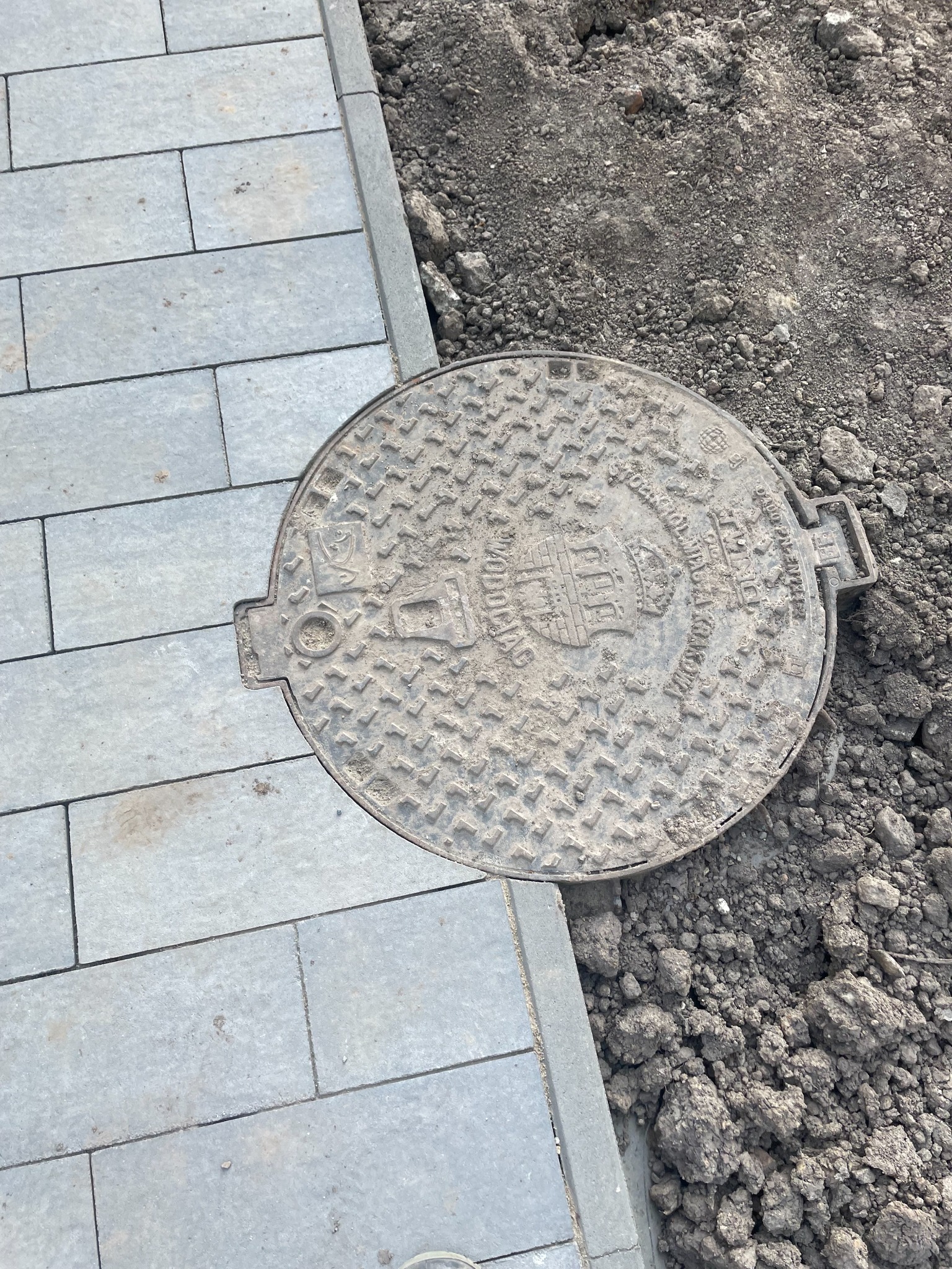 Szara kostka brukowa ułożona obok ziemi z żeliwną studzienką kanalizacyjną. Wykończona obrzeżami. Detal z bliska.