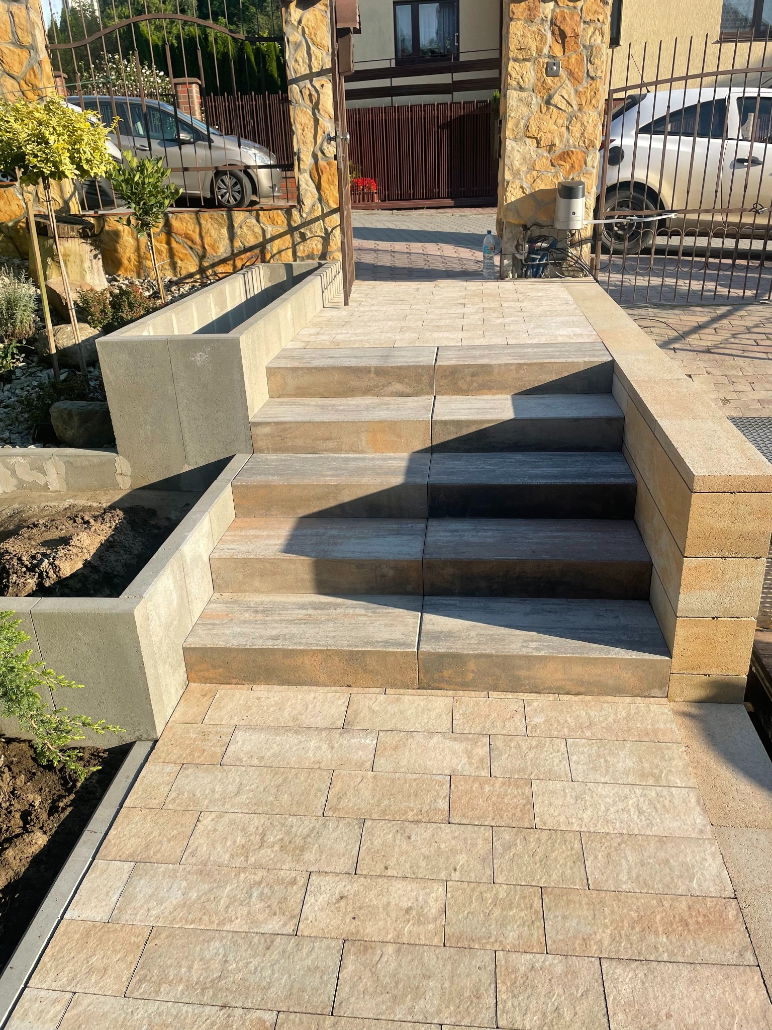 Schody z kostki brukowej prowadzące do bramy wjazdowej, z elementami małej architektury i rabatami kwiatowymi w tle. Ujęcie w słoneczny dzień.