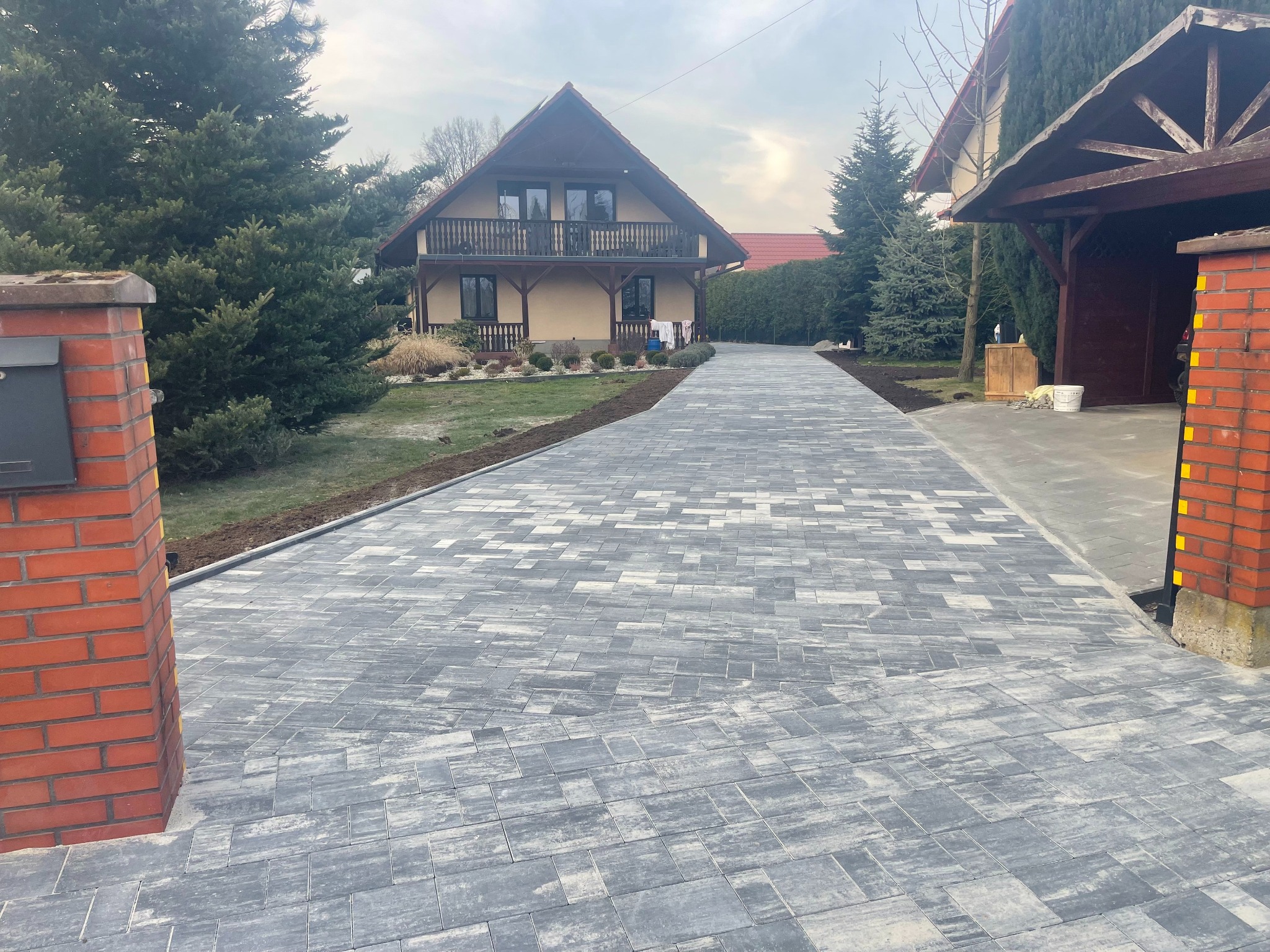 Nowa, szara kostka brukowa prowadzi do domu z balkonem. Świeżo ułożona nawierzchnia, widoczne obrzeża. Czerwona cegła w słupku przy wjeździe.