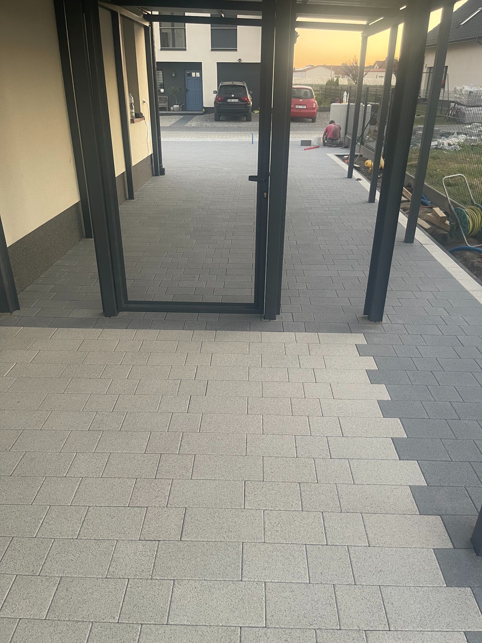 Ułożona kostka brukowa w dwóch odcieniach szarości pod zadaszeniem, z widocznym fragmentem układania i pracującą osobą w tle, w otoczeniu domów.