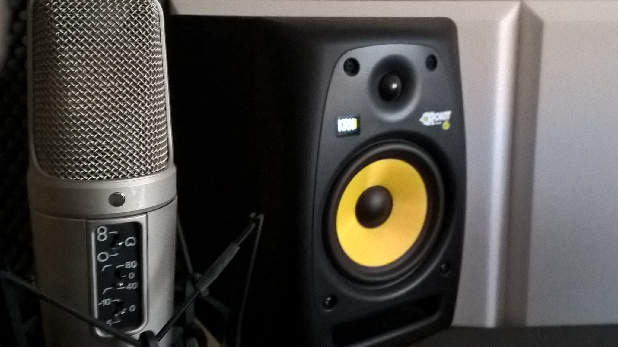 Mikrofon pojemnościowy z regulacją czułości obok czarnego monitora studyjnego z żółtą membraną na tle akustycznej ściany.