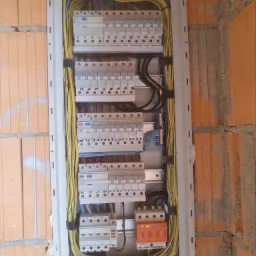 Rozdzielnica elektryczna z widocznymi przewodami, zamontowana na ścianie z cegły dziurawki, zabezpieczona uchwytami montażowymi.