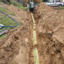 Wykop w ziemi z widocznymi rurami w żółtej folii zabezpieczającej, przygotowanymi do zasypania, na tle samochodu dostawczego z logo firmy instalacyjnej.
