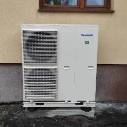 Zewnętrzna jednostka pompy ciepła Panasonic T-Cap 12kW, zamontowana na betonowej podstawie przy ścianie budynku o beżowej elewacji, widoczne okno z drewnianą ramą, teren zabezpieczony czarną folią.