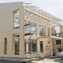 Surowa drewniana konstrukcja domu w trakcie budowy, widoczne rusztowania i dźwig samochodowy w tle.