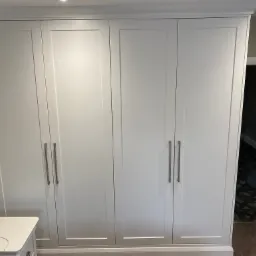 Biała, czterodrzwiowa szafa z prostymi, pionowymi uchwytami, zamontowana w pokoju we Wrocławiu. Obok szafy widoczny fragment białej szafki nocnej. W tle, przez otwarte drzwi, widać fragment podłogi...