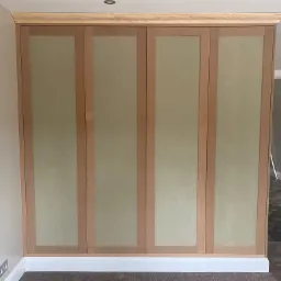 Czterodrzwiowa szafa wnękowa z surowego drewna i wypełnieniami z płyty MDF, w trakcie montażu, widoczne wykończenia sufitowe i podłogowe, z otwartymi drzwiami do sąsiedniego pokoju.