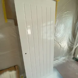 Świeżo pomalowane na biało drzwi wewnętrzne z pionowymi panelami, wiszące w warsztacie z folią ochronną na ścianach, widoczne ślady farby na podłodze.