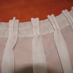 Jasnoszara, lniana zasłona z marszczeniami na taśmie, zbliżenie na górną krawędź i szlufki, widoczne delikatne prześwity materiału.