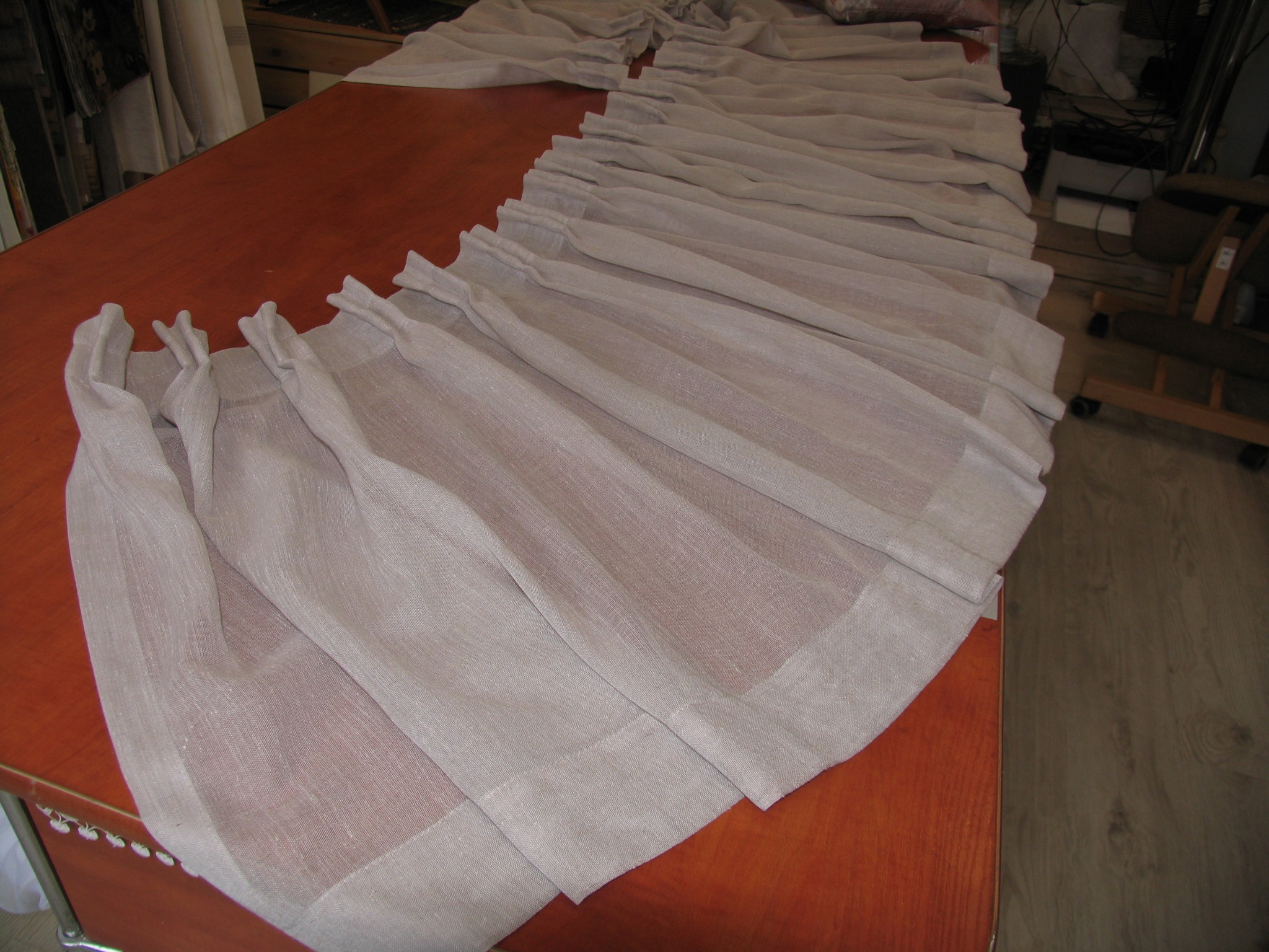 Fragment uszytej, beżowej zasłony z lnu z tunelem na karnisz, leżącej na brązowym stole.